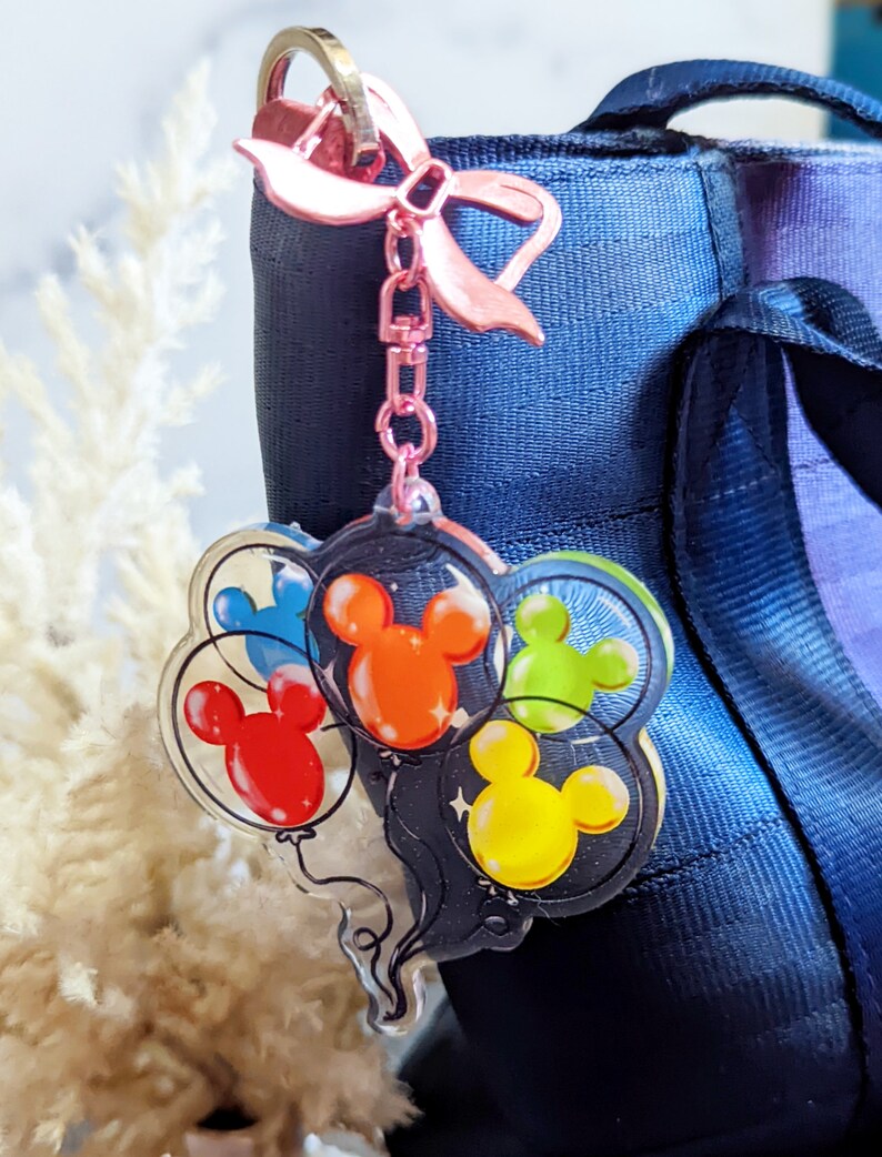Puede incluir: Un llavero de acr&iacute;lico transparente con un lazo rosa y una cadena. El llavero presenta un grupo de globos de Mickey Mouse en rojo, naranja, amarillo, verde y azul.
