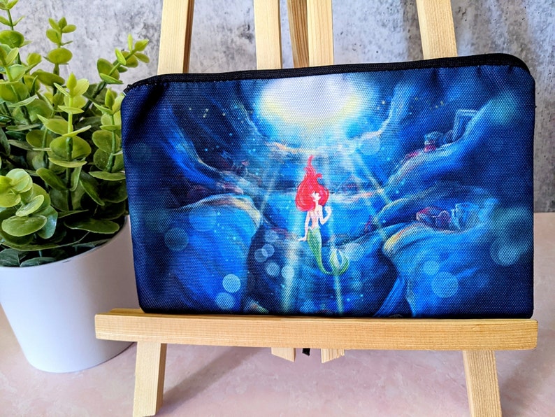 K&ouml;nnte beinhalten: Eine schwarze Rei&szlig;verschlusstasche mit einer farbenfrohen Illustration von Arielle aus Arielle, die Meerjungfrau. Die Illustration zeigt Arielle, wie sie mit einem blauen und gr&uuml;nen Schwanz und roten Haaren im Meer schwimmt. Der Hintergrund ist dunkelblau mit hellblauen Blasen.