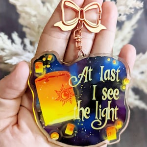 May include: A clear acrylic keychain with a gold-colored bow-shaped clasp. The keychain features a colorful design of a floating lantern with the sun symbol and the text "At Last I see the light" against a starry night sky.