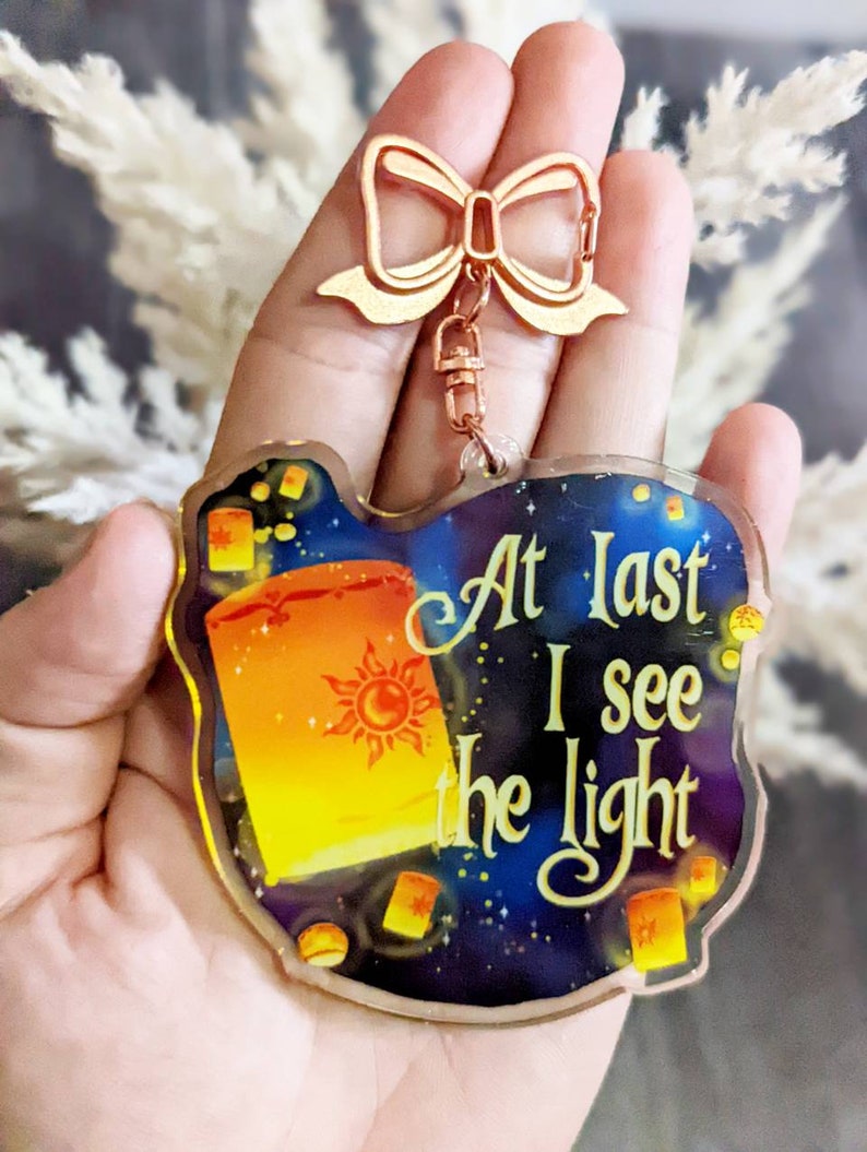 May include: A clear acrylic keychain with a gold-colored bow-shaped clasp. The keychain features a colorful design of a floating lantern with the sun symbol and the text "At Last I see the light" against a starry night sky.