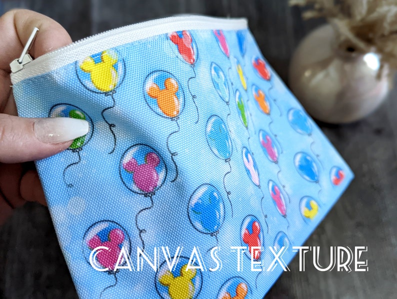 K&ouml;nnte beinhalten: Eine blaue Canvas-Rei&szlig;verschlusstasche mit einem wei&szlig;en Rei&szlig;verschluss. Die Tasche hat ein sich wiederholendes Muster aus bunten Luftballons mit Mickey-Maus-Ohren. Der Text "CANVAS TEXTURE" ist auf der Unterseite der Tasche aufgedruckt.