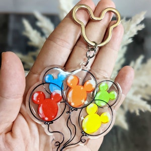 Puede incluir: Un llavero transparente con cinco globos coloridos con forma de cabeza de Mickey Mouse. Los globos son rojo, naranja, amarillo, verde y azul. El llavero tiene un anillo con forma de cabeza de Mickey Mouse.