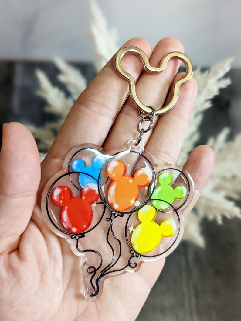 Puede incluir: Un llavero transparente con cinco globos coloridos con forma de cabeza de Mickey Mouse. Los globos son rojo, naranja, amarillo, verde y azul. El llavero tiene un anillo con forma de cabeza de Mickey Mouse.