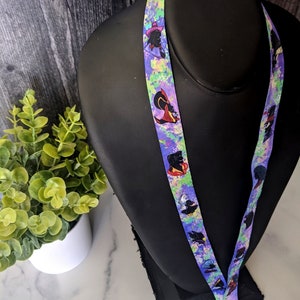 K&ouml;nnte beinhalten: Ein lilafarbenes Lanyard mit einem bunten Spritzlack-Design und wiederholten Bildern einer Cartoon-Figur mit einem lila Hut und schwarzen Haaren.