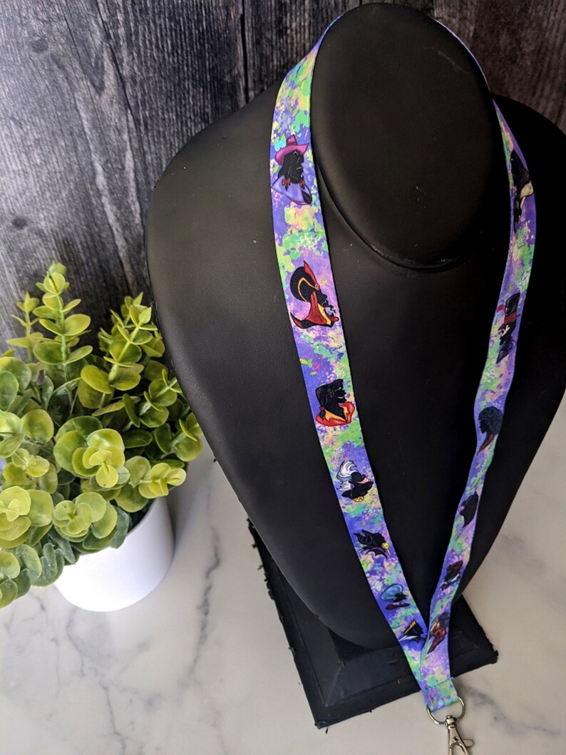 K&ouml;nnte beinhalten: Ein lilafarbenes Lanyard mit einem bunten Spritzlack-Design und wiederholten Bildern einer Cartoon-Figur mit einem lila Hut und schwarzen Haaren.