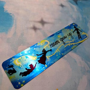 May include: A blue metal bookmark with a silhouette of Peter Pan and Tinkerbell flying with the text "never grow up" in white lettering.