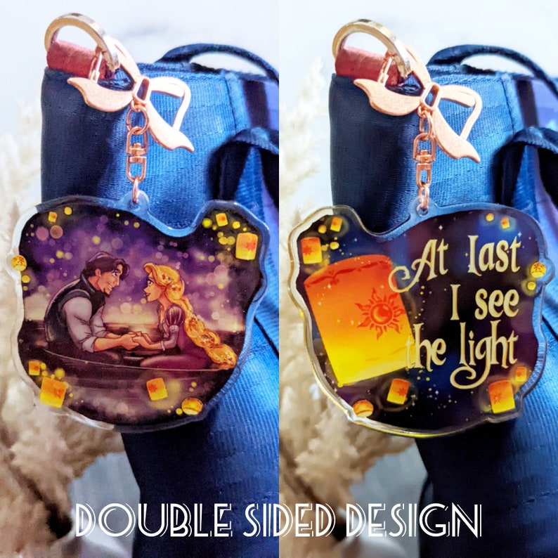 May include: Double-sided acrylic keychain featuring a romantic scene with a couple in a boat and the text "At last I see the light." The keychain has a rose gold bow and clasp. The design includes floating lanterns and a starry night sky.