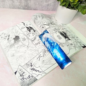 May include: A blue and white bookmark with a manga character and a starry sky design. The bookmark is placed on top of an open manga book.