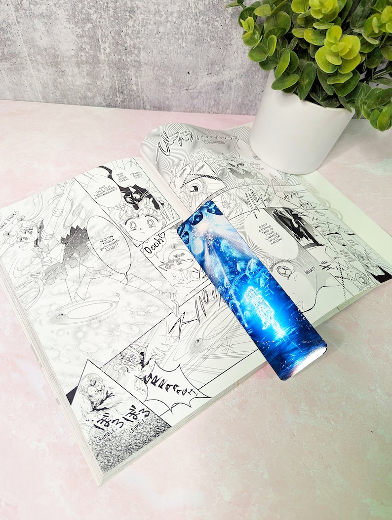 May include: A blue and white bookmark with a manga character and a starry sky design. The bookmark is placed on top of an open manga book.