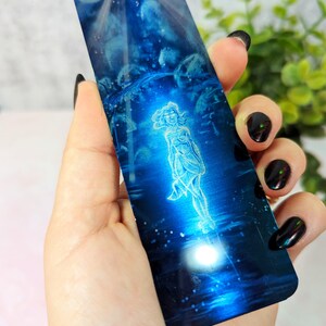 May include: A blue metal bookmark with a white image of a woman standing in a field of stars. The bookmark is shiny and reflective.