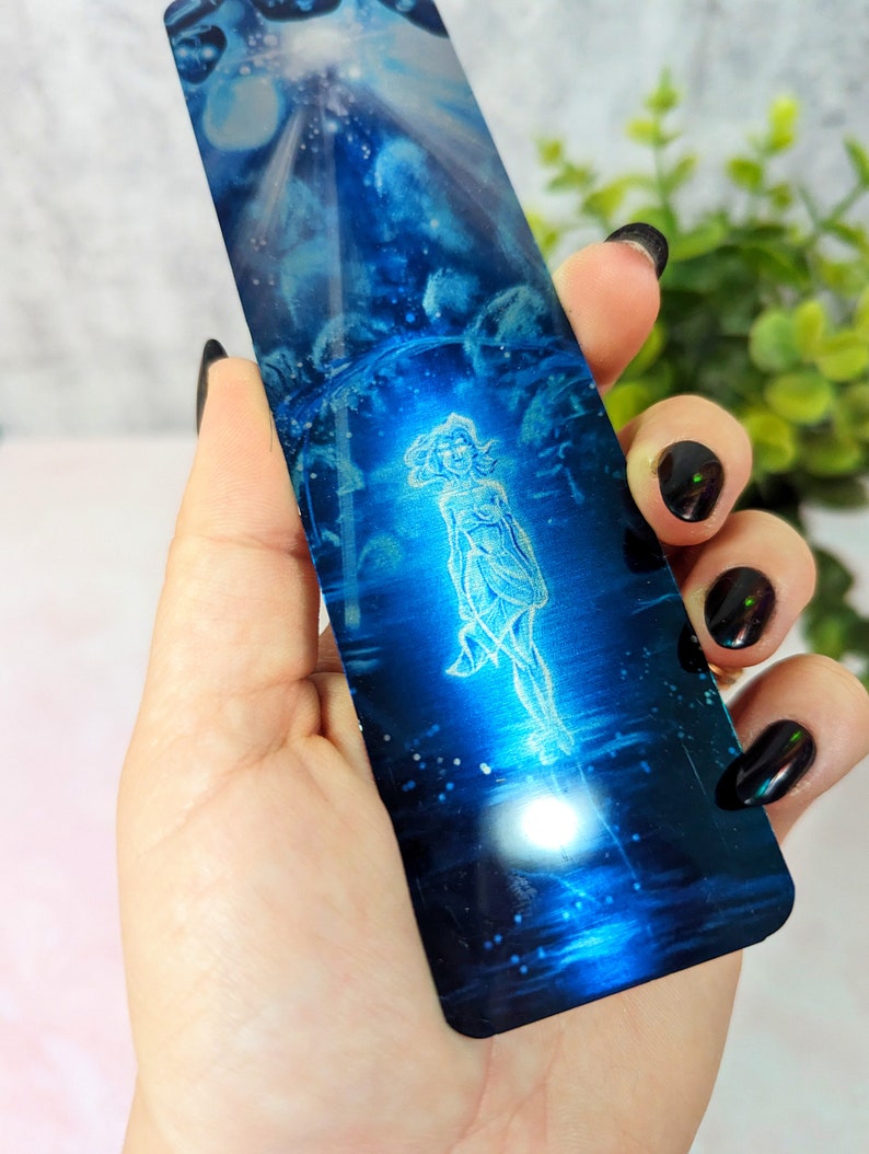 May include: A blue metal bookmark with a white image of a woman standing in a field of stars. The bookmark is shiny and reflective.
