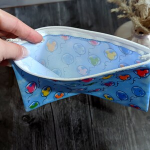 K&ouml;nnte beinhalten: Eine blaue Rei&szlig;verschlusstasche mit einem wei&szlig;en Rei&szlig;verschluss und einem wei&szlig;en Futter. Die Tasche ist mit einem Muster aus bunten Ballons mit Mickey-Maus-K&ouml;pfen verziert.