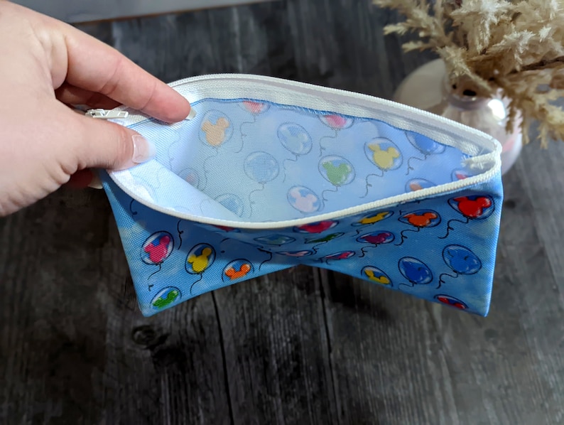 K&ouml;nnte beinhalten: Eine blaue Rei&szlig;verschlusstasche mit einem wei&szlig;en Rei&szlig;verschluss und einem wei&szlig;en Futter. Die Tasche ist mit einem Muster aus bunten Ballons mit Mickey-Maus-K&ouml;pfen verziert.