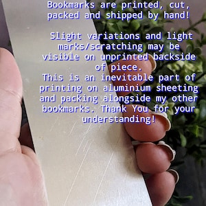 May include: Close-up of a silver bookmark with a hand holding it. The bookmark has a message that reads "*Please Note! Bookmarks are printed, cut, packed and shipped by hand! Slight variations and light marks/scratching may be visible on unprinted backside of piece. This is an inevitable part of printing on aluminium sheeting and packing alongside my other bookmarks. Thank You for your understanding!"