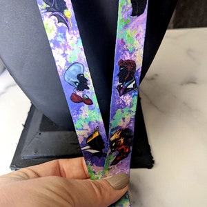 K&ouml;nnte beinhalten: Ein lila und gr&uuml;nes Lanyard mit einem aquarellartigen Design. Das Lanyard zeigt verschiedene Comicfiguren, darunter einen Wolf, eine Frau mit einer Krone und eine Frau mit blauen Haaren.