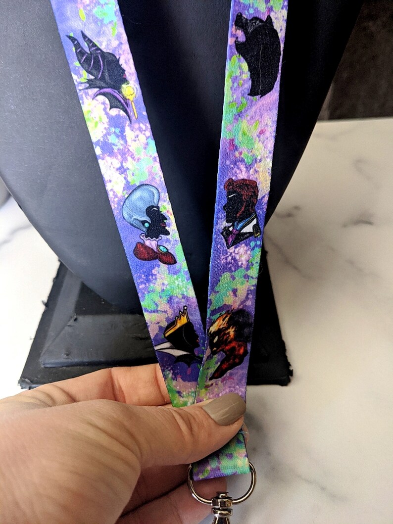 K&ouml;nnte beinhalten: Ein lila und gr&uuml;nes Lanyard mit einem aquarellartigen Design. Das Lanyard zeigt verschiedene Comicfiguren, darunter einen Wolf, eine Frau mit einer Krone und eine Frau mit blauen Haaren.