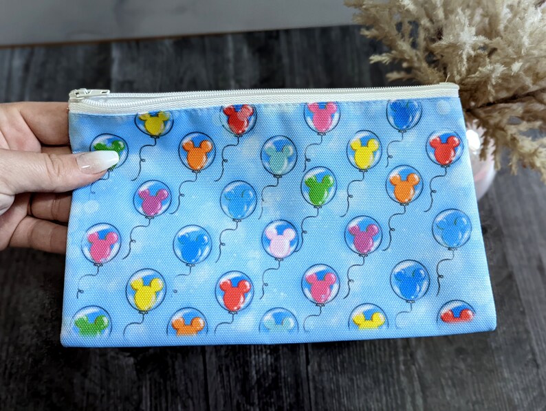 K&ouml;nnte beinhalten: Eine blaue Rei&szlig;verschlusstasche mit einem wei&szlig;en Rei&szlig;verschluss. Die Tasche ist mit einem Muster aus bunten Mickey-Mouse-K&ouml;pfen in Ballons verziert.