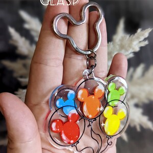 Puede incluir: Un llavero de Mickey Mouse plateado con un dije de cinco cabezas de Mickey Mouse de colores dentro de globos transparentes.