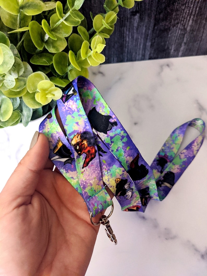 K&ouml;nnte beinhalten: Ein lilafarbenes Lanyard mit einem bunten Spritzlack-Design. Das Lanyard zeigt Comicfiguren mit schwarzen Umrissen. Das Lanyard hat einen silbernen Metallverschluss.