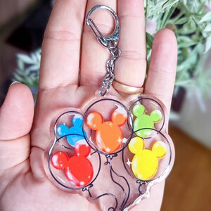 Puede incluir: Un llavero de plata con un dije que presenta cinco globos de Mickey Mouse de colores. Los globos son rojo, naranja, amarillo, verde y azul.