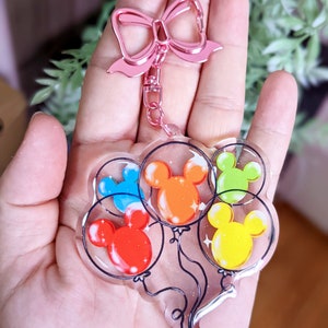 Puede incluir: Un llavero con un lazo rosa y un dije de cinco globos coloridos con orejas de Mickey Mouse. Los globos son transparentes y tienen brillo en su interior.