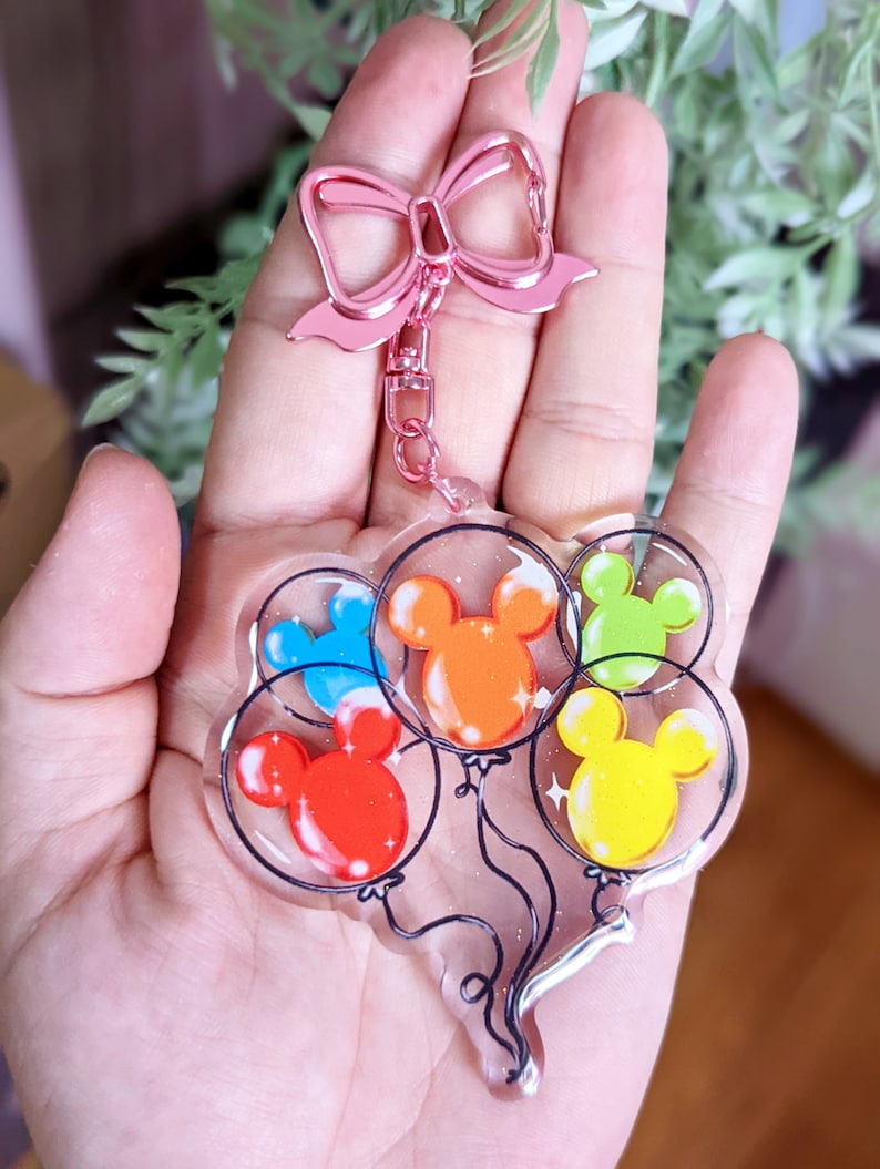 Puede incluir: Un llavero con un lazo rosa y un dije de cinco globos coloridos con orejas de Mickey Mouse. Los globos son transparentes y tienen brillo en su interior.