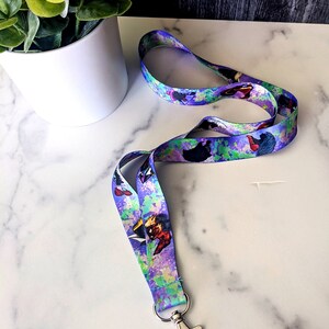 K&ouml;nnte beinhalten: Ein lila, blaues und gr&uuml;nes Lanyard mit einem aquarellartigen Design, das Comicfiguren zeigt. Das Lanyard hat einen silbernen Metallclip.