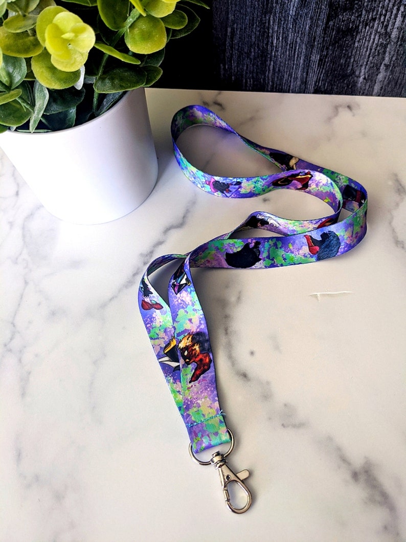 K&ouml;nnte beinhalten: Ein lila, blaues und gr&uuml;nes Lanyard mit einem aquarellartigen Design, das Comicfiguren zeigt. Das Lanyard hat einen silbernen Metallclip.