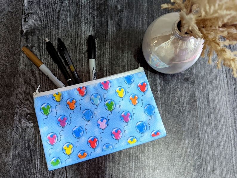 K&ouml;nnte beinhalten: Eine blaue Stiftebox mit Rei&szlig;verschluss. Die Box ist mit einem Muster aus bunten Ballons mit Mickey Mouse-Ohren verziert. Die Box ist mit Stiften und Markern gef&uuml;llt.