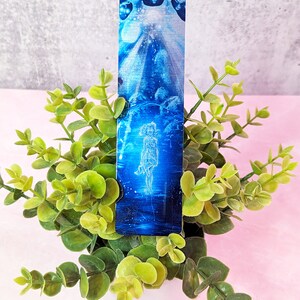 May include: A blue and white bookmark with a woman standing in a field of flowers. The bookmark is made of metal and has a shiny finish.