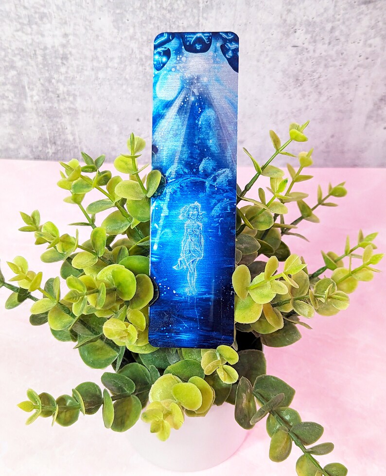 May include: A blue and white bookmark with a woman standing in a field of flowers. The bookmark is made of metal and has a shiny finish.