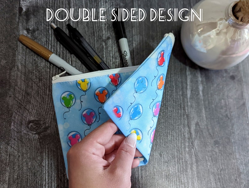 K&ouml;nnte beinhalten: Eine blaue Rei&szlig;verschlusstasche mit einem Mickey Mouse Ballon-Druck. Die Tasche ist ge&ouml;ffnet und zeigt das Innere, das ebenfalls mit Mickey Mouse Ballons bedruckt ist. Der Text "DOUBLE SIDED DESIGN" ist oben im Bild sichtbar.