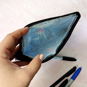 K&ouml;nnte beinhalten: Eine schwarze Rei&szlig;verschlusstasche mit einer durchsichtigen Plastikverkleidung. Die Verkleidung hat ein buntes Unterwassermotiv. Die Tasche ist offen und eine Hand h&auml;lt sie.