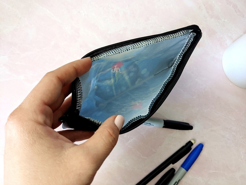 K&ouml;nnte beinhalten: Eine schwarze Rei&szlig;verschlusstasche mit einer durchsichtigen Plastikverkleidung. Die Verkleidung hat ein buntes Unterwassermotiv. Die Tasche ist offen und eine Hand h&auml;lt sie.