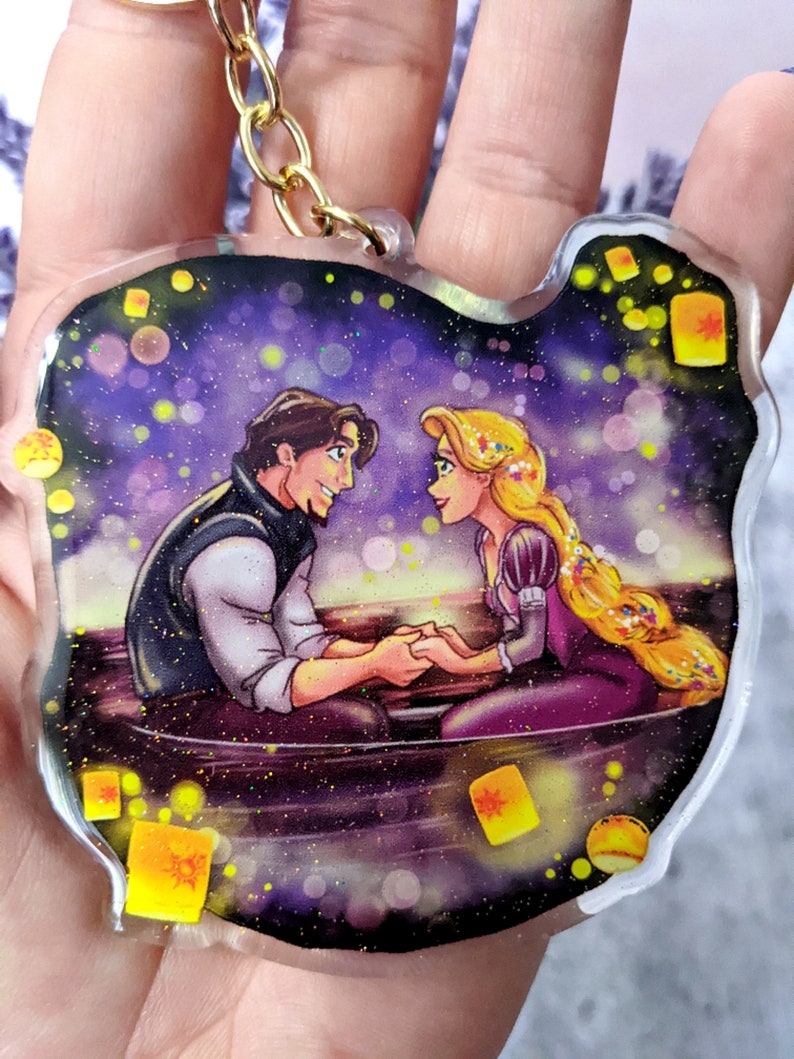 May include: A clear, cat-shaped keychain featuring a romantic scene with a man and woman in a boat, surrounded by floating lanterns. The artwork has a glittery effect and a gold-colored chain.