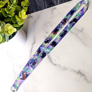 K&ouml;nnte beinhalten: Ein lilafarbenes Lanyard mit einem farbenfrohen Aquarelldesign, das verschiedene Comicfiguren zeigt. Das Lanyard hat an einem Ende einen silbernen Clip.