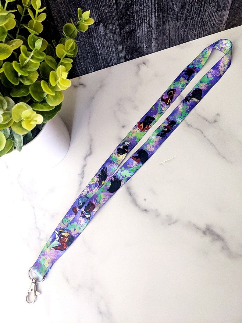 K&ouml;nnte beinhalten: Ein lilafarbenes Lanyard mit einem farbenfrohen Aquarelldesign, das verschiedene Comicfiguren zeigt. Das Lanyard hat an einem Ende einen silbernen Clip.