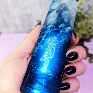 May include: A blue and silver bookmark with a glowing figure of a woman standing in a pool of water. The background is a dark blue with a light blue sky and stars.