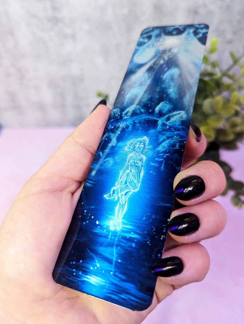 May include: A blue and silver bookmark with a glowing figure of a woman standing in a pool of water. The background is a dark blue with a light blue sky and stars.