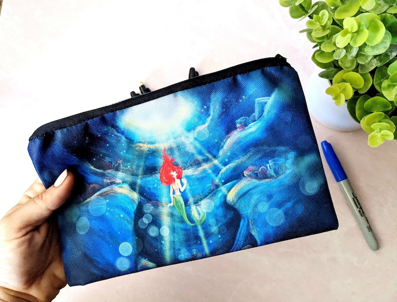 K&ouml;nnte beinhalten: Eine schwarze Canvas-Rei&szlig;verschlusstasche mit einer farbenfrohen Illustration von Arielle, der kleinen Meerjungfrau, die unter Wasser schwimmt. Die Illustration zeigt eine blaue, gr&uuml;ne und violette Ozeanszene mit Blasen und Licht, das von oben nach unten scheint.