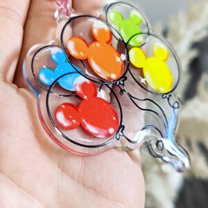 Puede incluir: Un llavero de resina transparente con un acabado de epoxi brillante que presenta cinco globos con forma de cabeza de Mickey Mouse en diferentes colores: rojo, naranja, amarillo, verde y azul.
