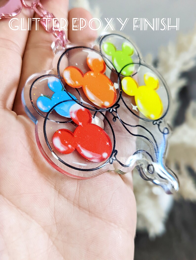 Puede incluir: Un llavero de resina transparente con un acabado de epoxi brillante que presenta cinco globos con forma de cabeza de Mickey Mouse en diferentes colores: rojo, naranja, amarillo, verde y azul.
