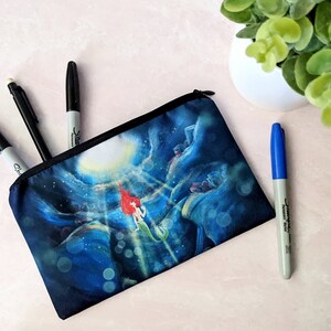 K&ouml;nnte beinhalten: Eine schwarze Rei&szlig;verschlusstasche mit einer blauen und wei&szlig;en Illustration von Arielle aus Arielle, die Meerjungfrau, die nach oben zu einer Lichtquelle schwimmt. Die Tasche liegt auf einer rosa Oberfl&auml;che mit drei schwarzen Markern und einem blauen Marker daneben.