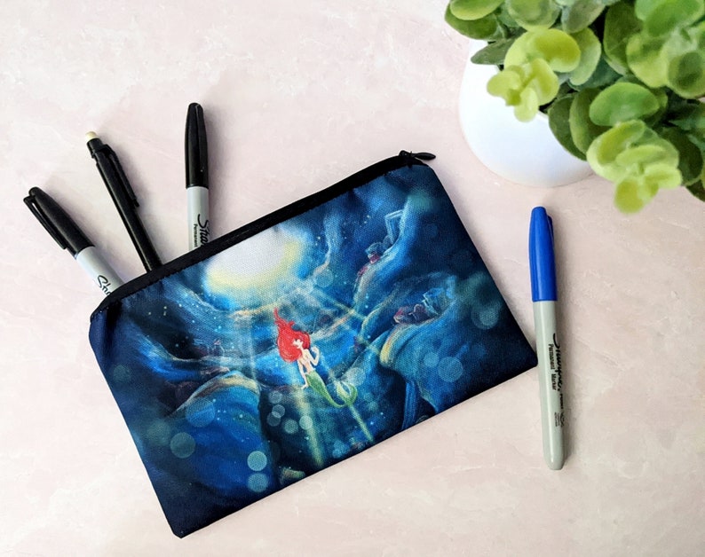 K&ouml;nnte beinhalten: Eine schwarze Rei&szlig;verschlusstasche mit einer blauen und wei&szlig;en Illustration von Arielle aus Arielle, die Meerjungfrau, die nach oben zu einer Lichtquelle schwimmt. Die Tasche liegt auf einer rosa Oberfl&auml;che mit drei schwarzen Markern und einem blauen Marker daneben.