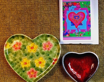 Puede incluir: Una colección de artículos en forma de corazón: un plato de cerámica verde con diseños florales, un cuenco rojo pequeño y una pila de tarjetas de felicitación con una ilustración temática de corazón. Los artículos están dispuestos sobre una superficie marrón texturizada.