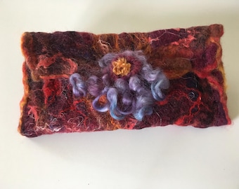 May include: A rectangular, handmade felt clutch in shades of red, burgundy, and orange. A central floral embellishment features a golden center and swirling lavender and blue accents. The textured surface suggests a unique, artisanal design.