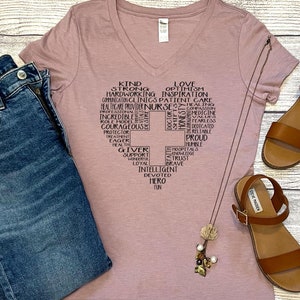 May include: A mauve v-neck t-shirt with a graphic design of a cross made up of words describing a nurse. The shirt is paired with blue jeans and brown sandals.