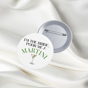 May include: White button with green text that reads "I'm the bride, pour me a martini". A martini glass with a green olive is illustrated below the text. The back of the button is silver.