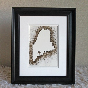 May include: A black framed picture with a white mat and a white silhouette of the state of Maine with two gold stars inside. The background is a brown speckled texture.