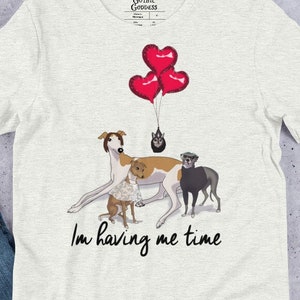 May include: Light gray t-shirt featuring a graphic of several dogs, including a greyhound, with red heart-shaped balloons. The text "I'm having me time" is printed below the dogs. The shirt has a crew neck and short sleeves.